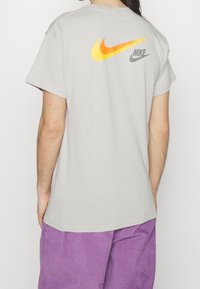 Person wearing light gray Nike t-shirt with two large orange and yellow swoosh logos on the upper back and purple corduroy pants.