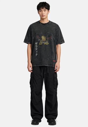 Jeune homme portant un t-shirt graphique noir avec un motif de dragon et un texte japonais, associé à un pantalon cargo noir et des baskets noires, debout face à nous.