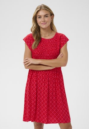 Žena s dlhými blond vlasmi, oblečená v červenom krátkom rukáve so vzorom, stojaca so založenými rukami a usmievajúca sa na jednoliatom pozadí.