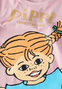 Maglietta di cotone rosa con una grande grafica colorata di un personaggio con capelli arancioni, lentiggini e una camicia blu. Sopra, in glitter dorato, c'è la scritta "PIPPI CALZALARGA."