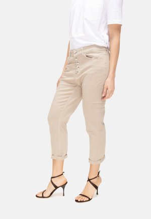 Donna che indossa pantaloni beige arrotolati con bottoni a vista, camicia bianca a maniche corte e sandali neri con tacco alto e cinturini, in piedi.
