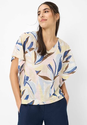 Vrouw met lang donker haar draagt een kortere mouwen, V-hals blouse met blauw, bruin en geel bladmotief, gecombineerd met donkerblauwe broek.