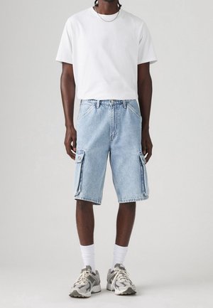 Homme portant un t-shirt blanc uni, un short cargo en jean bleu clair, des chaussettes blanches et des baskets grises et blanches, debout sur un fond clair.