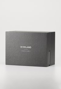 Čierna obdĺžniková obalová krabica s textúrovaným povrchom, na prednej strane s bielym textom "D1 MILANO" a geografickými súradnicami.