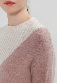 Pull en tricot avec un col côtelé blanc, panneaux diagonaux rose clair et beige. Texture douce avec une coupe décontractée et des détails subtils.
