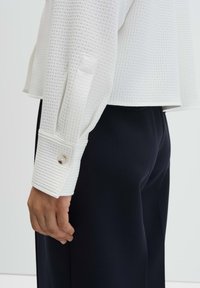 Top bianco a maniche lunghe con trama e design decorato, polsini con bottoni, abbinato a pantaloni aderenti blu navy, che mostrano una silhouette pulita.