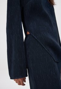 Top de manga larga plisado en color navy con tejido texturizado, que presenta un corte asimétrico y abertura lateral. Caída suave y silueta fluida.