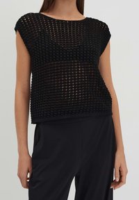 Musta teksturoitu neulepaita, jossa on leveä kaula-aukko ja hihattomat hihat. Rento malli on varustettu avoimella kudontakuviolla, ja se on paritettu tummien housujen kanssa.