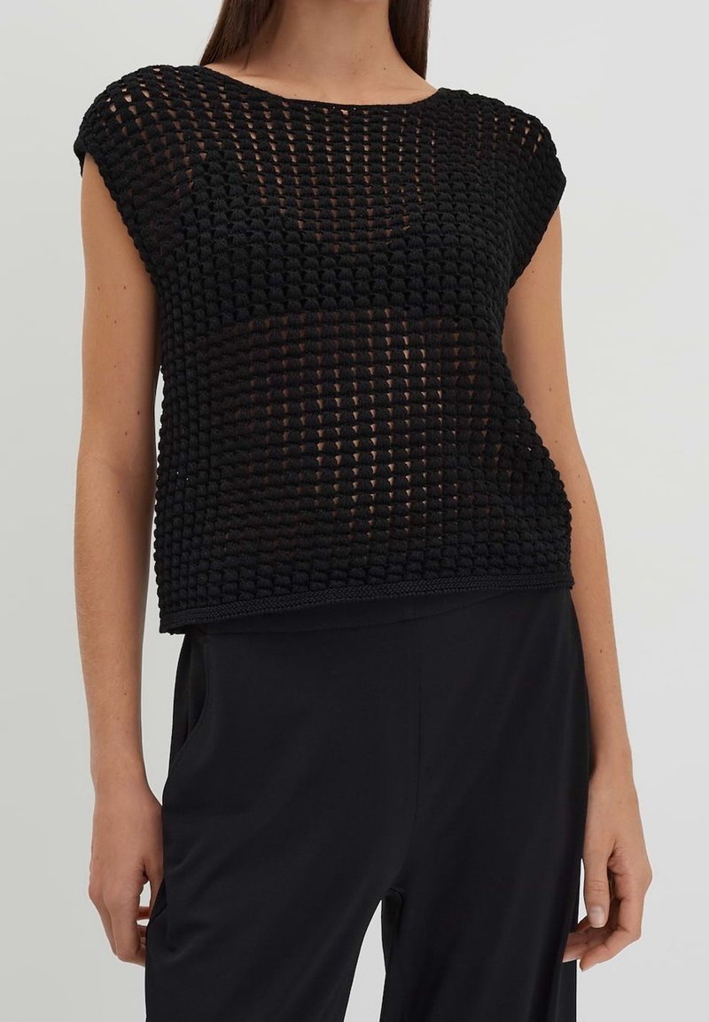 Musta teksturoitu neulepaita, jossa on leveä kaula-aukko ja hihattomat hihat. Rento malli on varustettu avoimella kudontakuviolla, ja se on paritettu tummien housujen kanssa.