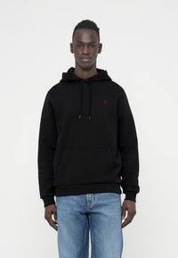 Zwarte hoodie met een voorzak, verstelbare trekkoorden en een klein rood logo. Gecombineerd met lichtblauwe spijkerbroek.