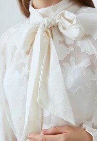 Blouse ivoire avec un motif floral texturé, un col haut et un grand nœud à la collerette. Fabriquée en tissu léger et transparent.