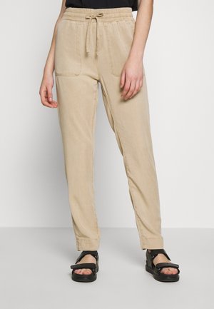 Pantalon beige à cordon avec poches avant, porté avec des sandales noires, montré de la taille aux pieds sur un fond uni.