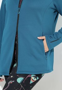 Tyrkysová fleecová bunda s černým zipem, bočními kapsami a detaily na manžetách, kombinovaná s legínami s potiskem v růžové a tyrkysové barvě.