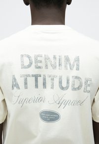 Κοντομάνικη κρεμ μπλούζα με μεγάλα, ξεθωριασμένα γκρι γράμματα στην πλάτη που γράφουν "DENIM ATTITUDE" και "Superior Apparel", με ένα λογότυπο στο κάτω μέρος.