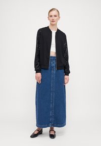 Black bomber jacket with sequin-embellished sleeves, paired with a long denim skirt. Model wears a white crop top and black flats.