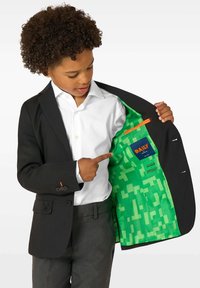 Poiss, kellel on seljas must ülikond ja valge särk, avab jakki, et näidata ere roheline mustriga vooder koos sisetaskute ja etiketiga.