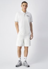 Camisa polo blanca con ribete negro y logo, combinada con pantalones cortos blancos que presentan el mismo logo. Zapatillas grises y calcetas blancas con logo completan el atuendo.