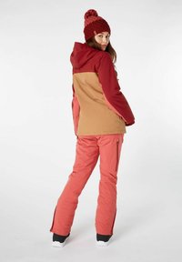 Ski-jack in bordeaux en tan, met een capuchon en verstelbare manchetten. Roze skibroek met zijritsen. Bruine gebreide muts met pompon.