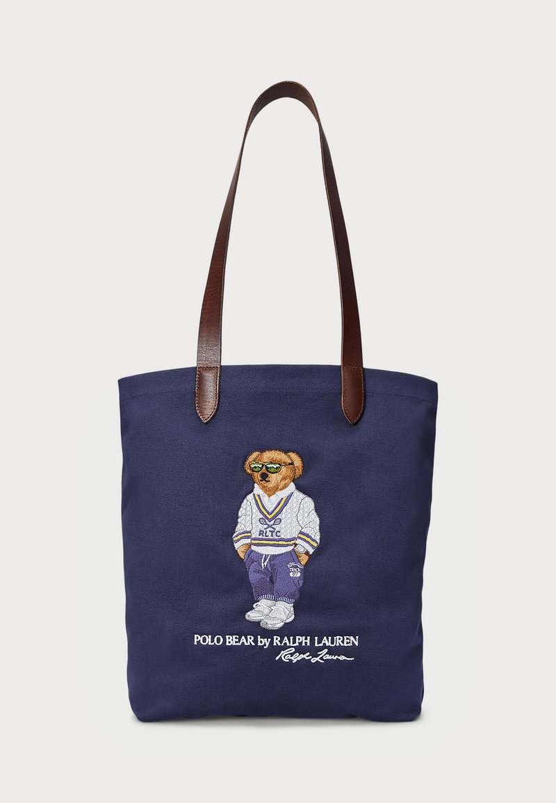 Sac fourre-tout en toile navy avec des poignées en cuir marron ; comporte un ours brodé en tenue de sport, portant des lunettes de soleil, avec du texte en dessous.