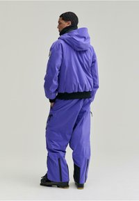 Lila szigetelt kabát kapucnival, rugalmas mandzsettával és oversize szabással, párosítva hozzá illő laza nadrággal, amely cipzárokkal és fekete díszítésekkel rendelkezik.