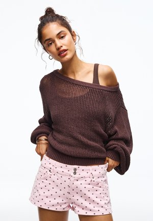 Young woman wears loose-knit off-shoulder brown sweater over tank and pink polka dot shorts, posing with hands in pockets.