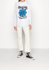 Langärmliges weißes Sweatshirt mit einem blauen Blumenmotiv und dem Text "Lasst uns gemeinsam wachsen!", kombiniert mit cremefarbenen Schlaghosen und schwarzen High-Top-Sneakern.