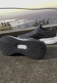Zapatilla deportiva negra con suela de goma texturizada, que presenta un diseño aerodinámico, parte superior de malla gris y tres líneas laterales sutiles.