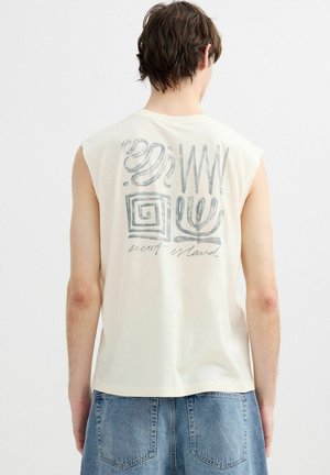 Hombre con camiseta blanca sin mangas con símbolos grises abstractos y el texto "secret island" en la espalda, combinado con vaqueros azules.