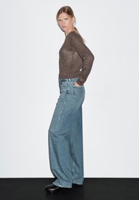 Hnědý texturovaný top s dlouhým rukávem v kombinaci s volnými světle modrými džínami se cvočky po stranách a černými dřeváky, prezentovaný na jednobarevném pozadí.