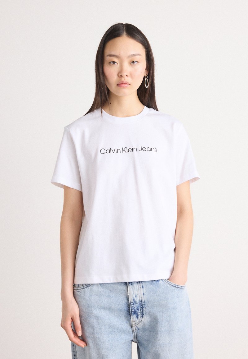 T-shirt in cotone bianco con collo rotondo e maniche corte. Sul davanti è presente la scritta "Calvin Klein Jeans" in testo nero. Abbinato a jeans azzurri chiari.