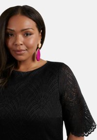 Černý krajkový top s texturovaným vzorem a vlnitými rukávy. Modelka má na sobě velké růžové a béžové náušnice. Hladké vlasy, neutrální líčení.