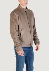 Giacca in suede marrone con colletto rialzato, chiusura con zip e due tasche laterali, abbinata a jeans blu. Tessuto liscio e design aderente.