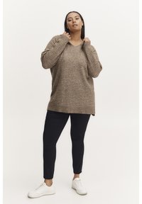 Pull beige en tricot avec un col en V et un cordon de serrage, associé à un pantalon slim bleu marine et des baskets blanches. Présente un motif texturé.