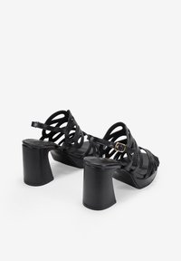 Black strappy high-heeled sandals with a chunky heel, featuring intertwined straps and a buckle detail at the ankle. Smooth texture.