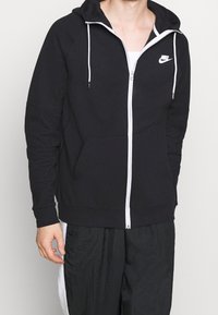 Svart zip-up hoodie gjort av mjukt tyg, med en vit dragkedja och logotyp, justerbar dragsko i huvan och framfickor.