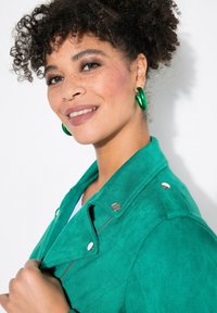 Giacca di suede verde con dettagli in metallo argentato, dotata di una zip asimmetrica e spalline. Modello che indossa orecchini a cerchio verdi coordinati.