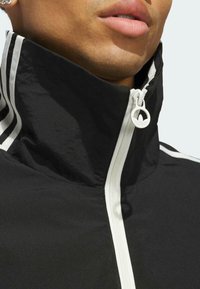 Schwarze Jacke mit hohem Kragen, weißem Reißverschluss und weißen Streifen an den Seiten. Das Material wirkt glatt mit einer matten Textur.