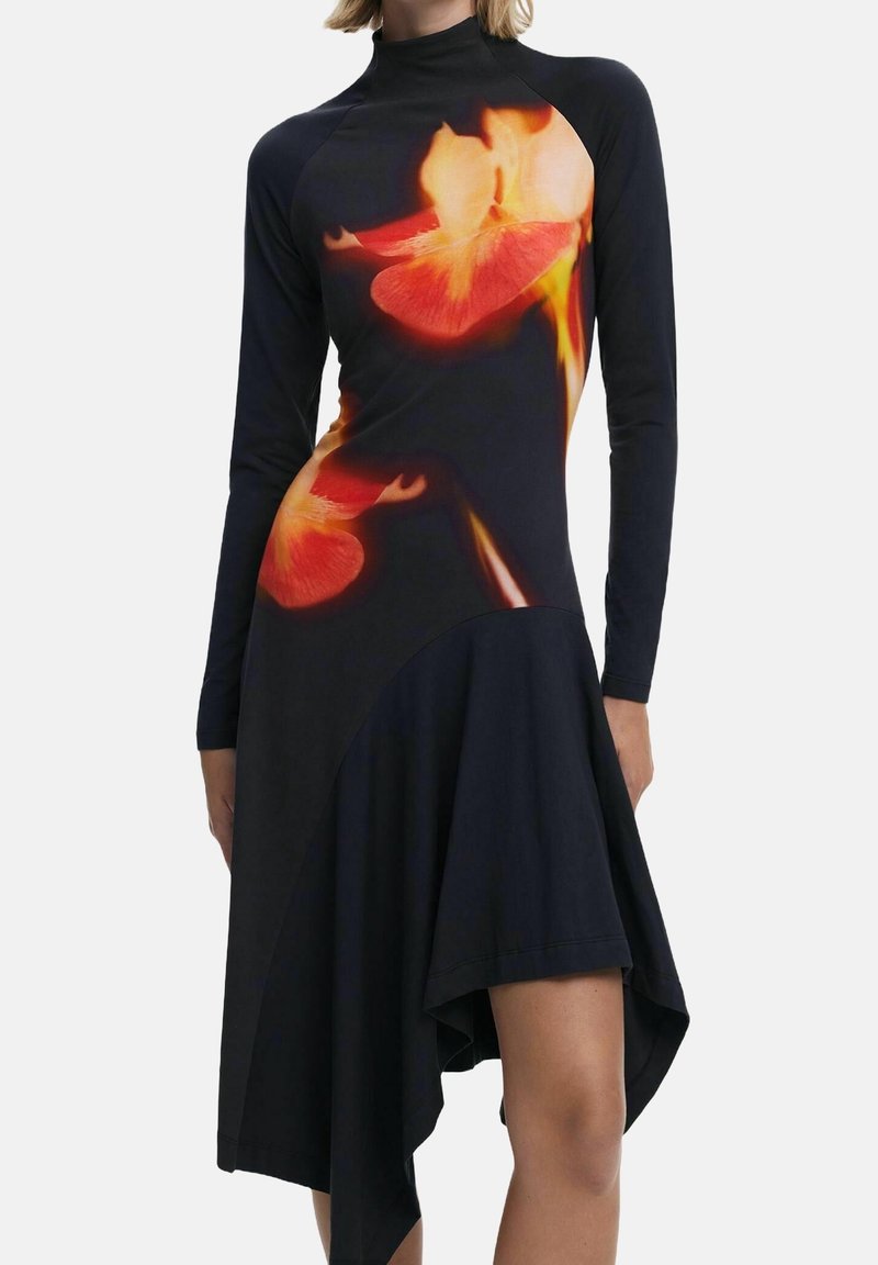 Femme portant une robe noire asymétrique à manches longues, ornée de grands motifs floraux orange vif et jaune sur les parties haute et moyenne.