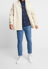 Béžová bavlněná bunda s límcem a předními kapsami, nošená přes tmavomodrou košili na knoflíky, zkombinována se světlými slim džínami a bílými teniskami.