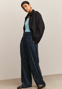 Veste noire surdimensionnée sur un débardeur bleu clair, associée à un pantalon en velours côtelé bleu foncé ample et des chaussures plates noires. Tissus lisses et texturés.