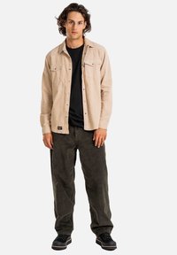 Camicia beige a maniche lunghe con due tasche sul petto, abbinata a pantaloni in velluto a coste verde scuro e scarpe nere con suola spessa.