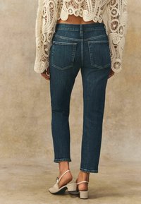 Vaqueros azul oscuro de corte slim, con un dobladillo ligeramente recortado y dos bolsillos traseros. Combinados con una blusa de punto color crema y tacones beige.