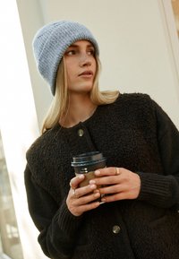 Zwarte gebreide cardigan met knoopdetails, gecombineerd met een lichtgrijze gebreide muts. Houdt een donkere beker met een kartonnen hoes vast.