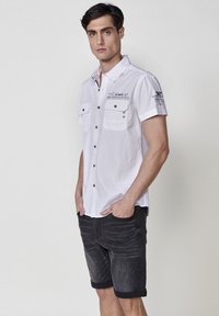 Camisa blanca de manga corta con botones y dos bolsillos en el pecho, botones negros y sutiles detalles en texto; combinada con pantalones cortos de mezclilla gris oscuro.