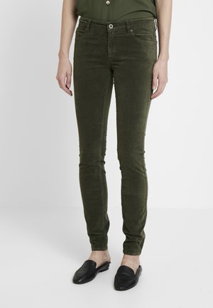 Frau trägt olivgrüne, figurbetonte Hose und schwarze Slipper, steht vor einem schlichten, hellen Hintergrund.