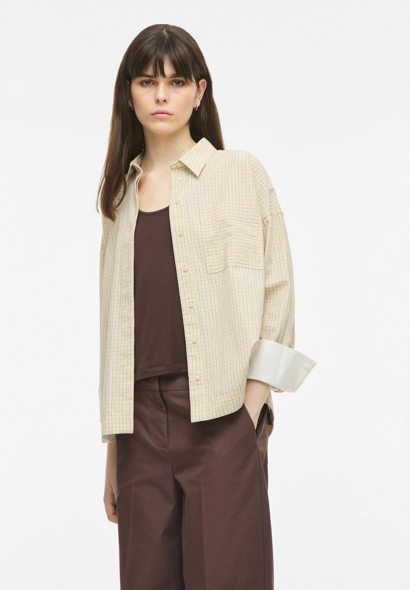 Jeune femme aux cheveux raides et foncés, portant une chemise beige à carreaux par-dessus un haut marron et un pantalon marron, debout avec une main dans la poche.