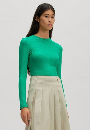 Woman with long dark hair wearing a fitted green ribbed long-sleeve top and high-waisted beige pleated skirt standing against a plain background.