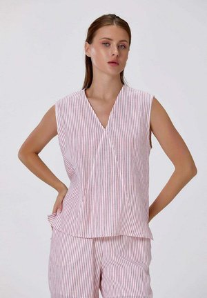 Femme portant un haut sans manches à rayures verticales roses et blanches et un pantalon assorti, posant avec les mains sur les hanches sur un fond uni.