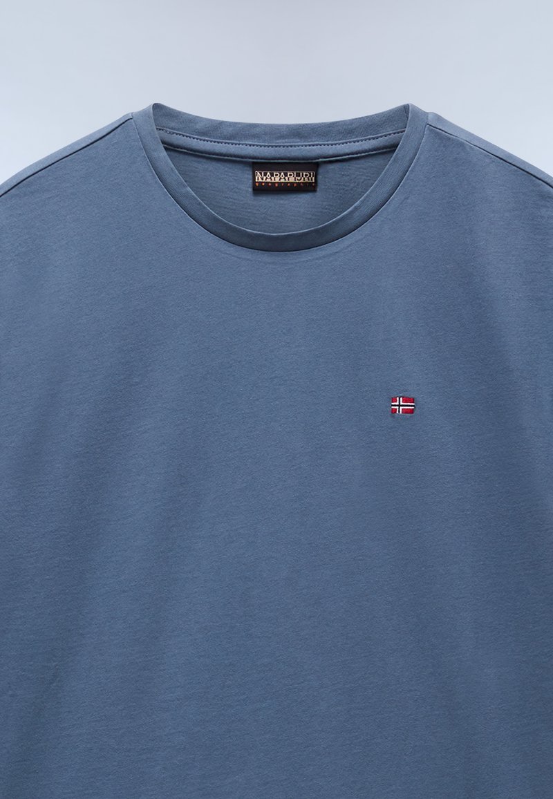 T-shirt in cotone di un blu chiaro morbido, con girocollo, maniche corte, che presenta una piccola bandiera norvegese ricamata in rosso e bianco sul petto.