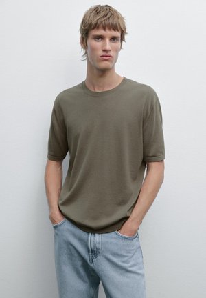 Ung mand med kort blond hår, iført en olivengrøn kortærmet skjorte og lyseblå jeans, står med hænderne i lommerne foran en hvid væg.